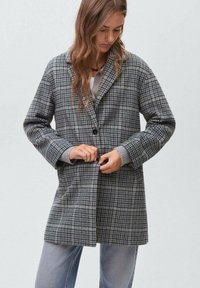 Gray plaid coat with a notch lapel, single button closure, and long sleeves, paired with a light gray sweater and denim jeans.