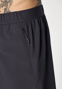 Nærbilde av svarte treningsshorts med teksturert stoff, elastisk midje, sidelomme med glidelås, og en tatovering delvis synlig på den nærliggende huden.