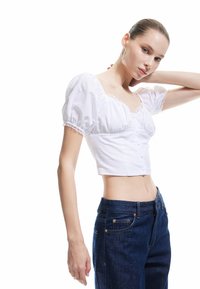 Weiße, kurz geschnittene Bluse mit Puffärmeln, Spitze und Knopfverschluss, kombiniert mit dunkelblauen, hoch taillierten Jeans mit goldenen Akzenten.
