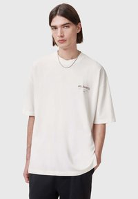 Camiseta de algodón blanca con un corte relajado, escote redondo, mangas cortas y un sutil logo de "A.L.L. SAINTS" en el pecho. Textura suave.