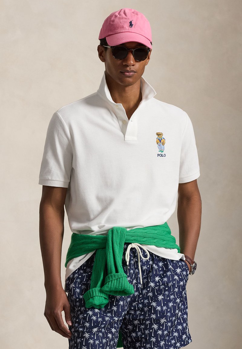 Mann iført hvit polo-skjorte med bjørnelogo, marineblå shorts med palmeprint, grønn genser knyttet rundt livet, rosa cap og solbriller.