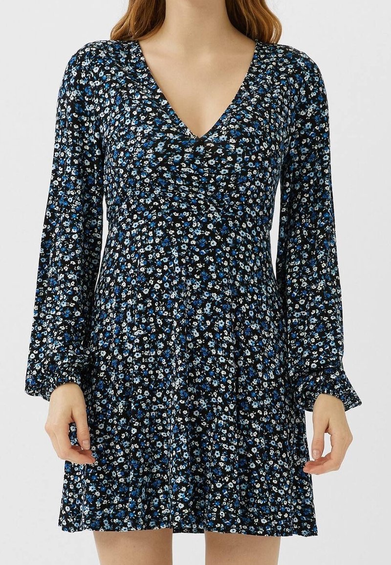 Femme portant une robe noire à manches longues à fleurs bleues et blanches petites avec un décolleté en V, debout avec les mains détendues sur les côtés.