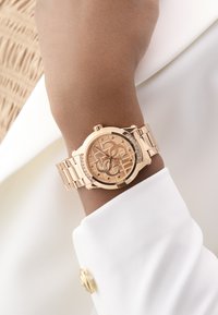 Roségoldene Armbanduhr mit transparentem Zifferblatt, verlinktem Armband und kleinen Strasssteinen. Getragen an einer Hand mit hellfarbigem Blusenärmel.