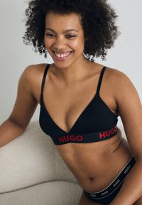 Zwarte driehoekige beha met rode "HUGO" logo band, verstelbare bandjes en een zachte stof; gecombineerd met bijpassende zwarte onderbroeken met logo band.
