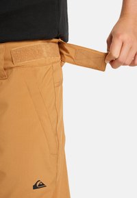 Hnedé snehové nohavice majú suchý zips v páse, hladký povrch, bočné vrecká a logo pri leme. Ruka uchopila nastavovač pásu.