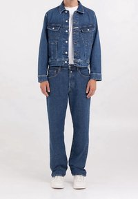 Jeansjacka med två framfickor, i kombination med matchande jeans, båda i en djup blå färg, och vita sneakers. Enkel vit T-shirt under.