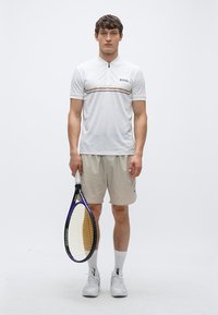 Λευκό αθλητικό πόλο μπλουζάκι με μισό φερμουάρ, με καφέ και μαύρη ρίγα. Κρατούνται ανοιχτό μπεζ σορτς και μοβ ρακέτα του τένις.