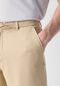 Pantalones beige texturizados con un ajuste relajado, que cuentan con un cinturón ajustable y bolsillos laterales. La superficie presenta un patrón tejido sutil.