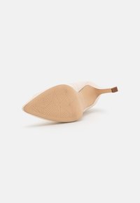 Chaussure à talon beige couchée sur le côté, montrant une semelle texturée avec des motifs de grip incurvés et une fine pointe de talon.