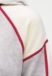 Κοντινό πλάνο πλεκτού πουλόβερ με κρέμ και ανοιχτό γκρι πάνελ, χωρισμένα από κόκκινες ραφές στον ώμο και το μανίκι.