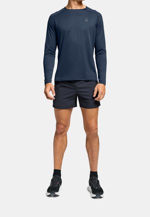 Mann steht in navyblauem Langarmshirt, schwarzen Sportshorts, schwarzen Socken und schwarzen Laufschuhen vor einem hellgrauen Hintergrund.
