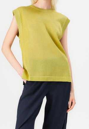 Ærmeløs lysegrøn strikket top med ribkanter, båret med mørke løse bukser, modellens venstre hånd i lommen.