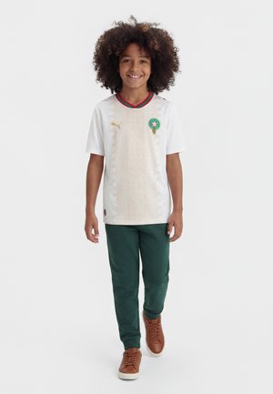 Παιδί με σγουρά μαλλιά χαμογελά, φοράει λευκή φανέλα ποδοσφαίρου με πράσινο και κόκκινο γιακά, σκούρο πράσινο παντελόνι και καφέ αθλητικά, προχωρώντας μπροστά.