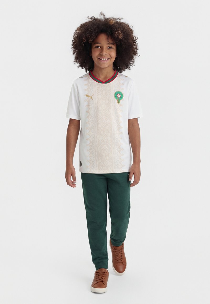 Παιδί με σγουρά μαλλιά χαμογελά, φοράει λευκή φανέλα ποδοσφαίρου με πράσινο και κόκκινο γιακά, σκούρο πράσινο παντελόνι και καφέ αθλητικά, προχωρώντας μπροστά.
