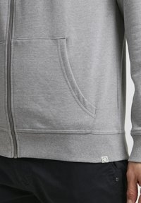 Haut à capuche zippé gris avec des poignets et un ourlet côtelés, des poches kangourou et une étiquette en tissu sur le côté inférieur. Mélange de coton doux et texturé.