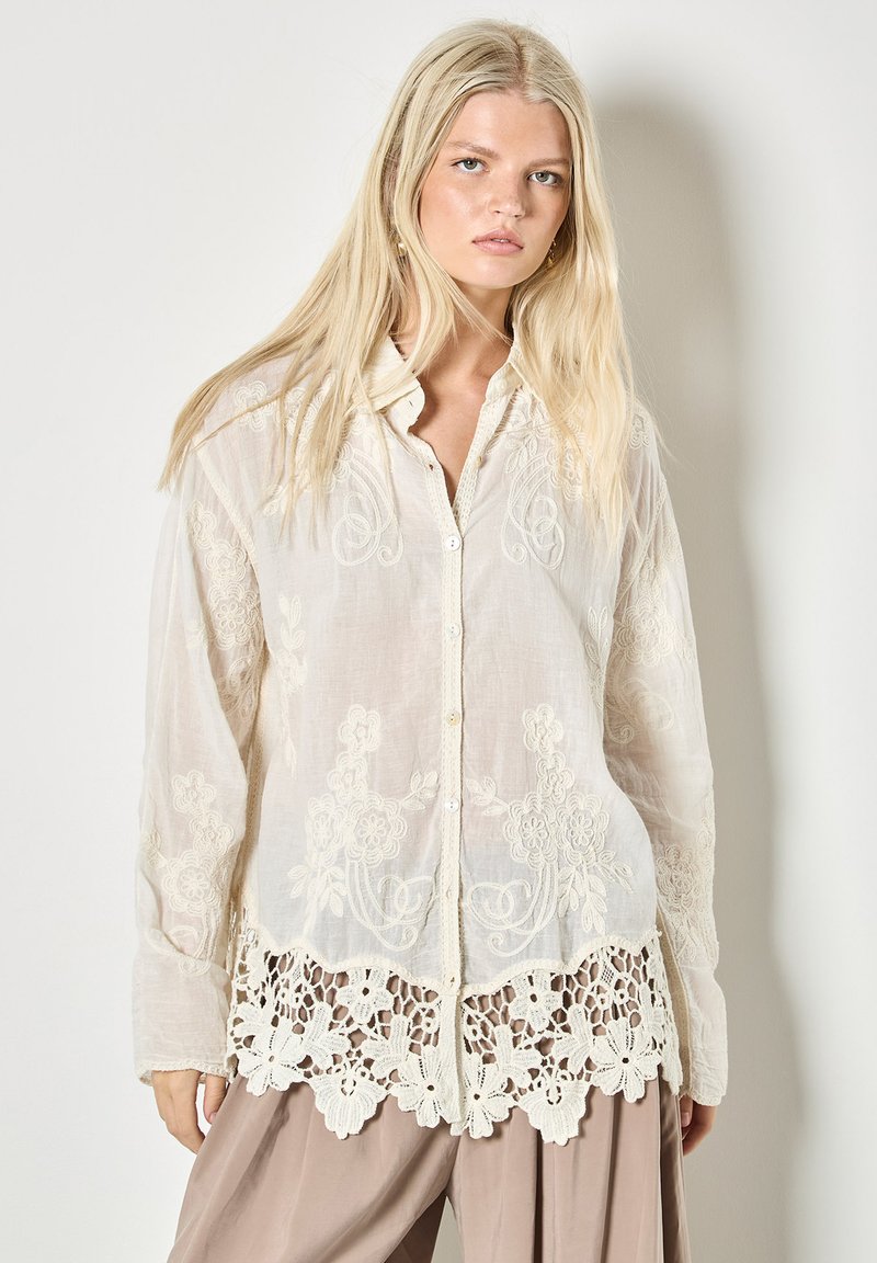 Femme blonde portant un chemisier blanc transparent brodé avec un ourlet en dentelle florale et un pantalon beige à jambes larges, se tenant devant un fond uni.
