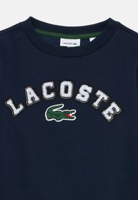 Navy zila crewneck džemperis ar ribotu apkakli, baltu izšūtu "LACOSTE" uzrakstu un zaļu krokodilu logotipu zem teksta.