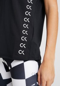 Zbliżenie osoby noszącej czarną bluzkę z pionowym rzędem białych logo „CK” oraz legginsy z czarno-białym geometrycznym wzorem i małym logo „CK”.