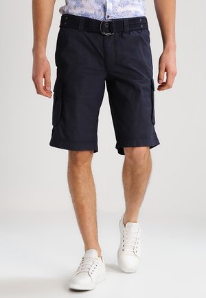 Mann trägt marineblaue Cargo-Shorts mit Seitentaschen, schwarzen Gürtel, weiße Sneaker und ein hellblau gemustertes Hemd, geht nach vorne.