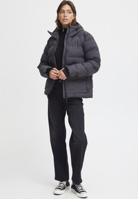 Giubbotto imbottito grigio scuro, con cappuccio, chiusura a zip frontale e polsini elasticizzati. Indossato sopra una maglia nera, abbinato a jeans neri e sneakers.