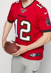 Punainen NFL Buccaneers -pelipaita mustilla ja valkoisilla yksityiskohdilla, jossa numero 12. Pelaajalla on käsissään ruskea jalkapallo ja hänellä on harmaat verkkarit.