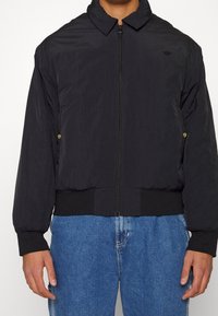 Černá lehká bomber bunda s stojáčkovým límcem, zipovým zapínáním a elastickými manžetami a lemem. Obsahuje dvě zlaté patentky na bocích.