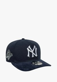 Puuvillainen tummansininen baseball-lippis, jossa on teksturoitu lippa, valkoinen kirjailtu "NY" -logo ja pienempi harmaa merkki sivussa.