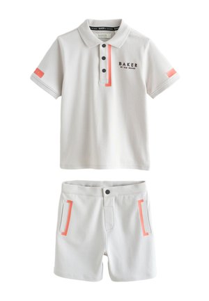 Dječak stoji u svijetlo sivoj polo majici i odgovarajućim kratkim hlačama, nosi bijele tenisice i čarape, na jednobojnoj svijetloj pozadini.