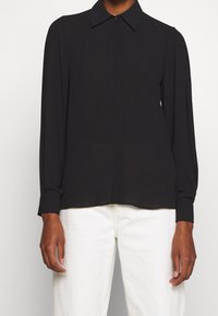 Camisa negra de manga larga con cuello puntiagudo, cierre frontal de botones y detalles plisados en los hombros. La tela tiene una textura suave.