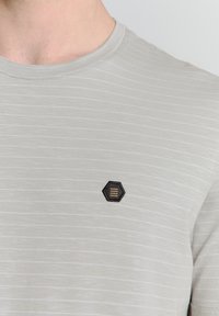 Lichtgrijze T-shirt met subtiele horizontale strepen, ronde halslijn en een hexagonaal geborduurd logo met drie horizontale lijnen.