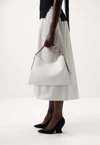 Sac à main en cuir blanc avec un bord supérieur incurvé, des anses noires et un accent métallique. Le modèle porte une jupe blanche fluide et des escarpins noirs à talons pointus.