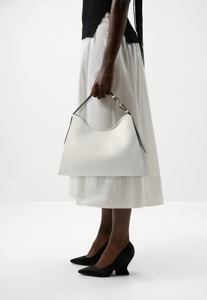 Weißes Lederhandtasche mit abgerundetem oberen Rand, schwarzen Trägern und einem metallischen Akzent. Das Modell trägt einen fließenden weißen Rock und schwarze spitze Absätze.