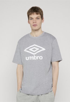 Junger Mann mit kurzen braunen Haaren, der ein graues Umbro-T-Shirt und passende graue Hosen vor einem schlichten hellen Hintergrund trägt.