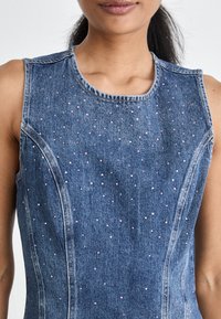 Denim ermeløs topp med rund hals, prydet med små rosa perler, med buede sømmer og en strukturert passform. Stoffet er mellomblå.