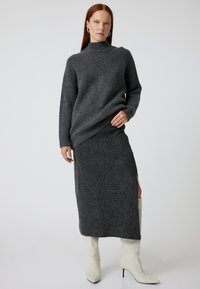 Pull en maille gris avec col montant, associé à une jupe maxi grise avec fente sur le côté. Stylisé avec des bottes à talons blanches.
