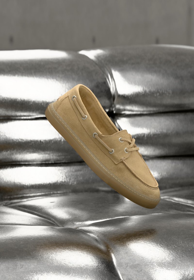 Μπεζ μοκασίνι από σουέντ με κορδόνια που αιωρείται πάνω από έναν αντανακλαστικό ασημένιο δερμάτινο καναπέ με κουμπιά.