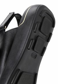 Sandale noire avec un dessus en cuir lisse et une semelle en caoutchouc texturée. Présente un léger accent doré le long du bord. Sangle ajustable incluse.