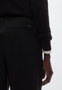 Zwarte broek met een vlakke achterzak, voorzien van een klein merkaccent. De stof is glad met een lichte glans en de tailleband is elastisch.