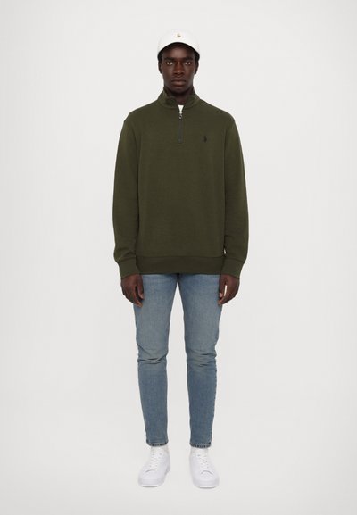 Mørkegrøn zip-up sweater med ribbede manchetter og kant, parret med lyseblå jeans og hvide sneakers, toppet med en hvid kasket.