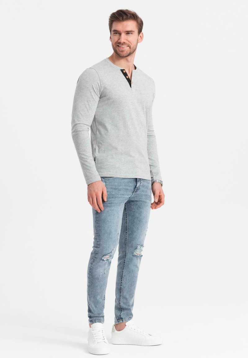 Camisa gris de manga larga con detalles de botones negros, combinada con jeans rasgados de color azul claro y zapatillas blancas, con un diseño ajustado.