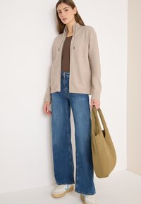 Beige Reißverschluss-Sweater mit hohem Kragen, kombiniert mit weiten blauen Jeans. Hält eine grüne Tasche und trägt weiße sowie hellblaue Sneakers.