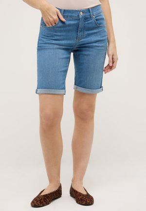 Mujer con shorts de mezclilla azul claro de longitud media con dobladillos enrollados y zapatos planos con estampado de leopardo, de pie contra un fondo neutro.