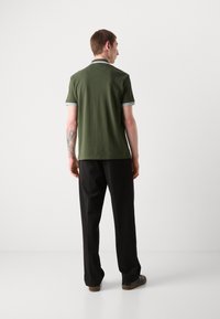 Kaki grønn polo skjorte med en subtil krage, vertikale striper på mansjettene; sammen med svarte bukser og brune sko. Enkel design og tekstur.