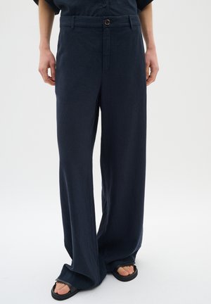 Persoon draagt donkerblauwe losse broek met knoopsluiting en zwarte sandalen op een witte achtergrond.