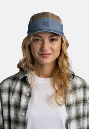 Jonge vrouw met golvend blond haar, die een blauw en beige pet, een wit T-shirt en een grijs-groen geruit overhemd draagt tegen een effen lichte achtergrond.