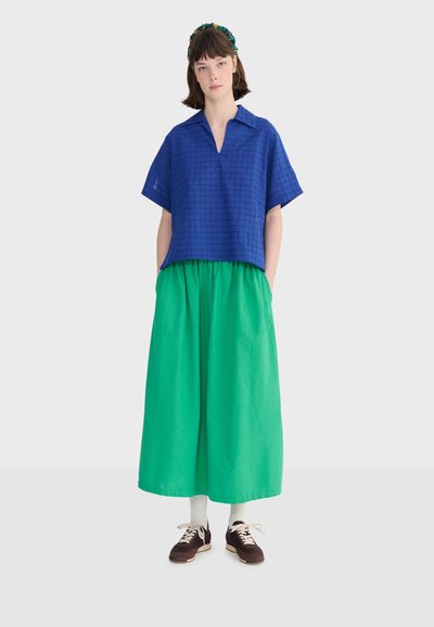 Femme portant un haut bleu à manches courtes et col en V, une jupe large verte vif, des chaussettes blanches, des baskets marron et un bandeau multicolore.