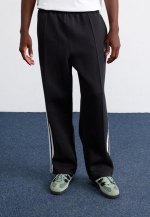 Black sweatpants with white side stripes, featuring an elastic waistband, worn with green sneakers on a blue carpeted surface.