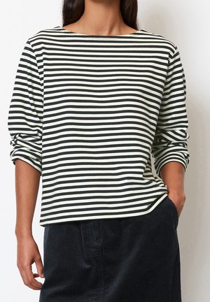 Mujer con una camiseta de rayas horizontales blancas y negras de manga larga con las mangas remangadas y pantalones de pana negros, con la mano en el bolsillo.