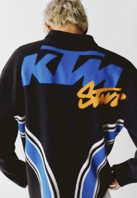 Czarny sweter dziewiarski z niebieskimi wzorami w zygzaki i pomarańczowym napisem. Białe akcenty podążają za krzywymi liniami wzdłuż boków. Wysoka konstrukcja kołnierza.