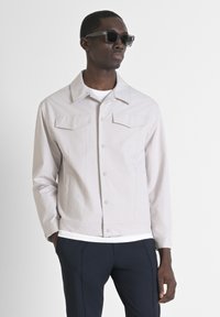 Lichtgrijze jas met knopen, twee borstzakken en een hoge kraag, gecombineerd met een wit shirt en marineblauwe broek.